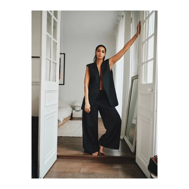 Fashionable woman in black vest and wide-leg pants poses barefoot in doorway with white French doors, bedroom visible in background.