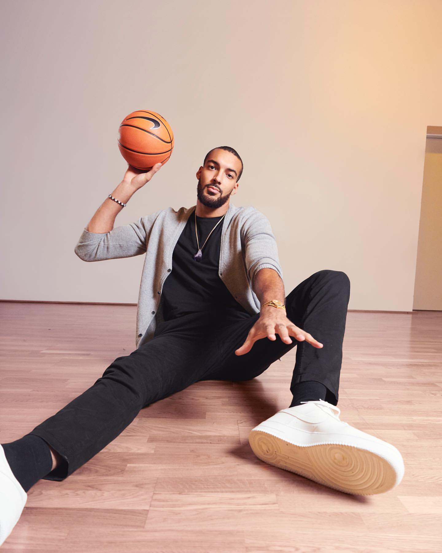 An adult male sits on a wood floor with a basketball in hand, wearing a gray cardigan, black t-shirt, black pants, and white shoes.