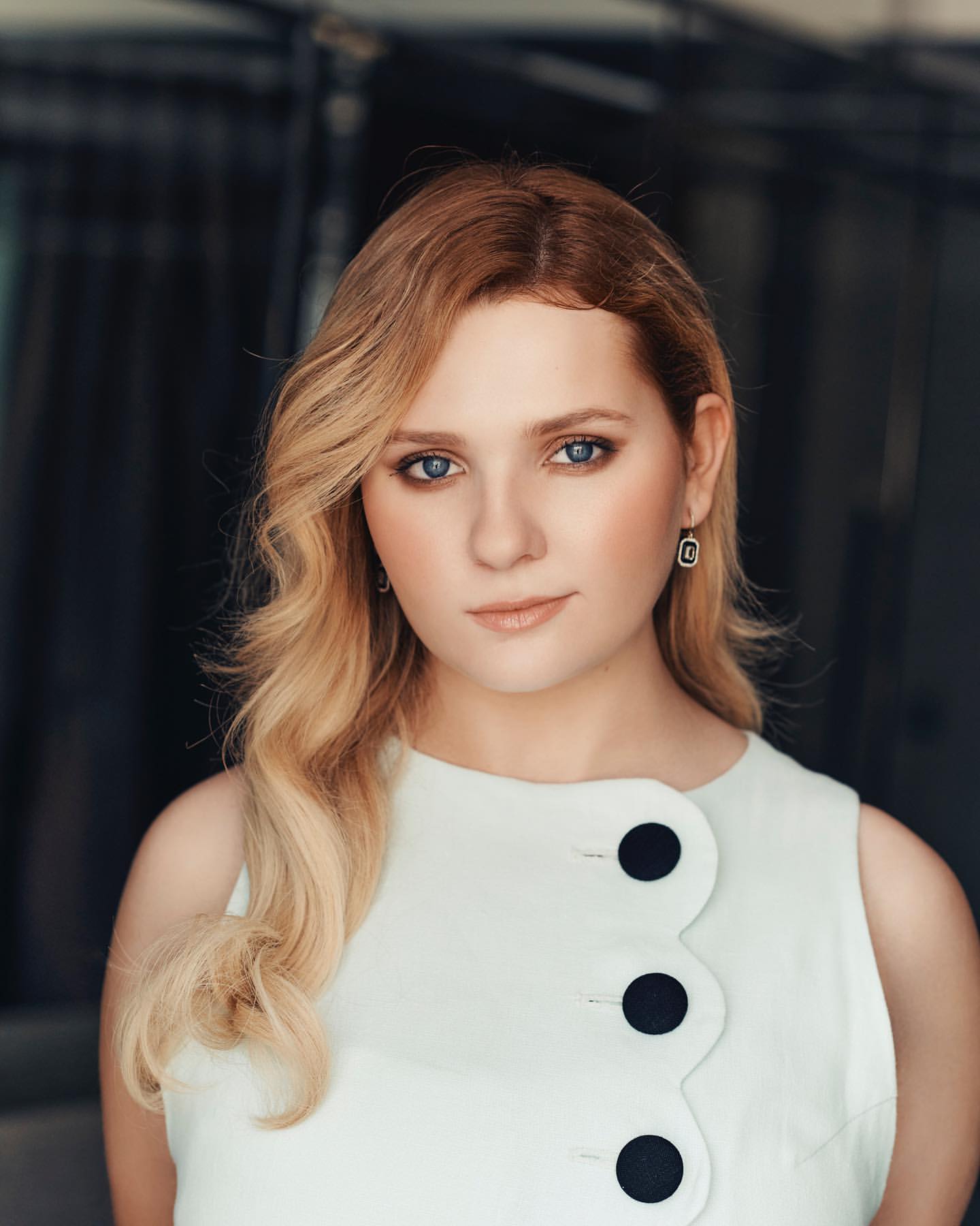 Portrait of a blond adult woman with blue eyes wearing a white sleeveless dress with large black buttons, posing indoors with a neutral expression.