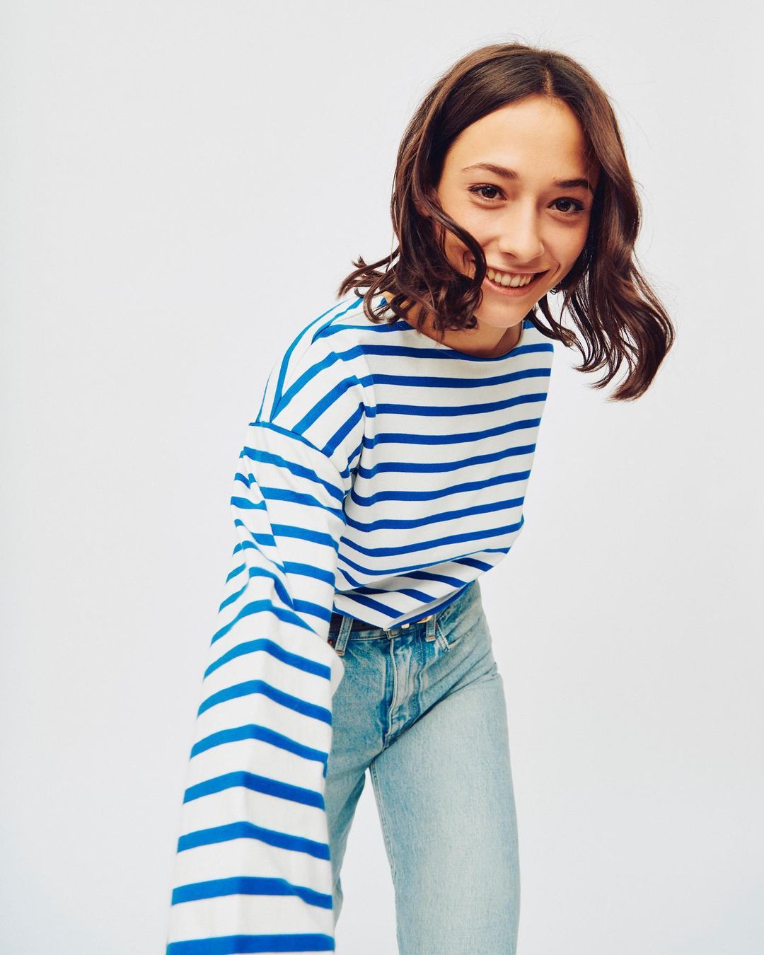 A smiling young woman with wavy brown hair wearing a blue and white striped shirt and light blue jeans poses against a white backdrop.