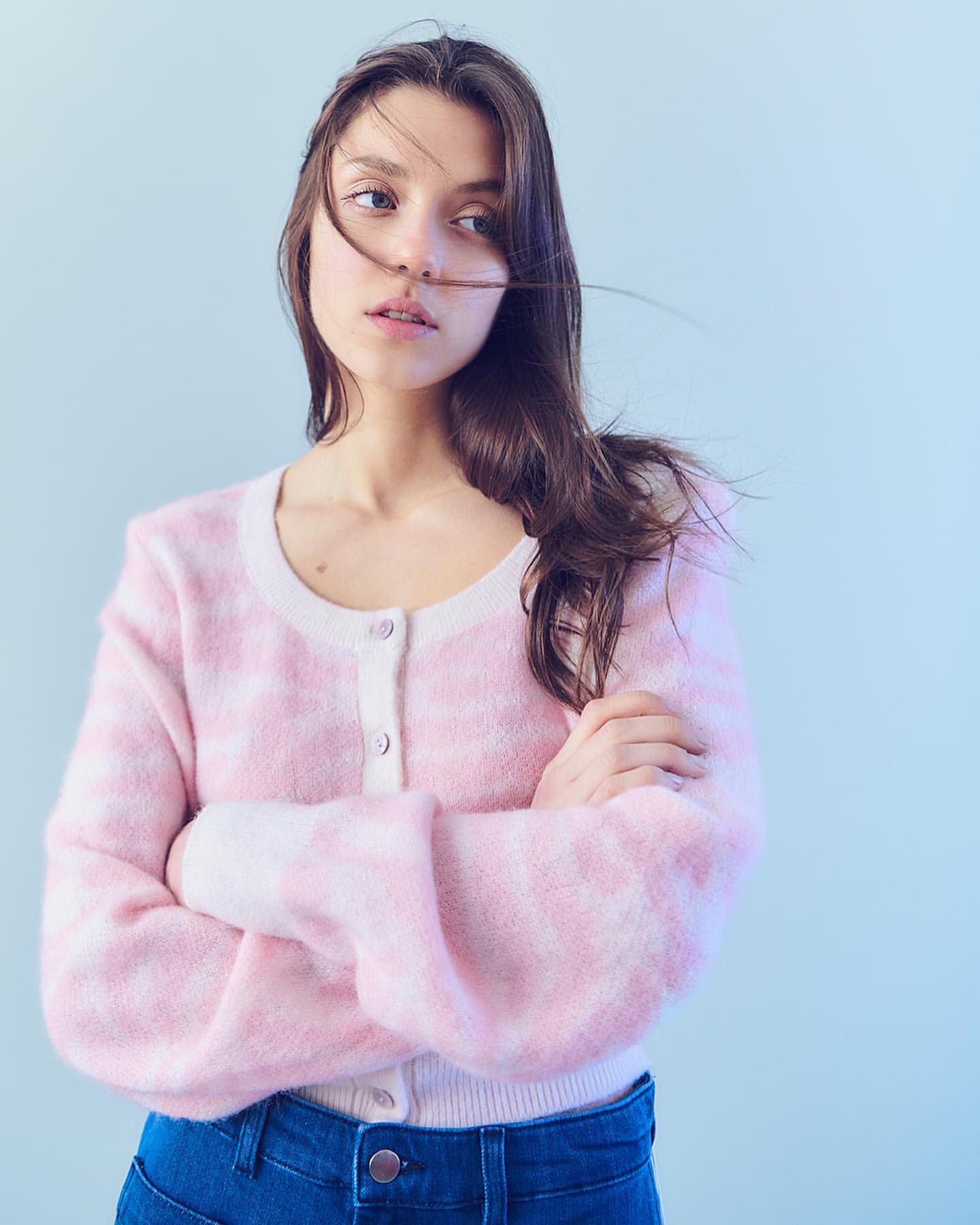A woman with brown hair and blue eyes poses indoors wearing a pink and white sweater and blue jeans against a light blue wall.