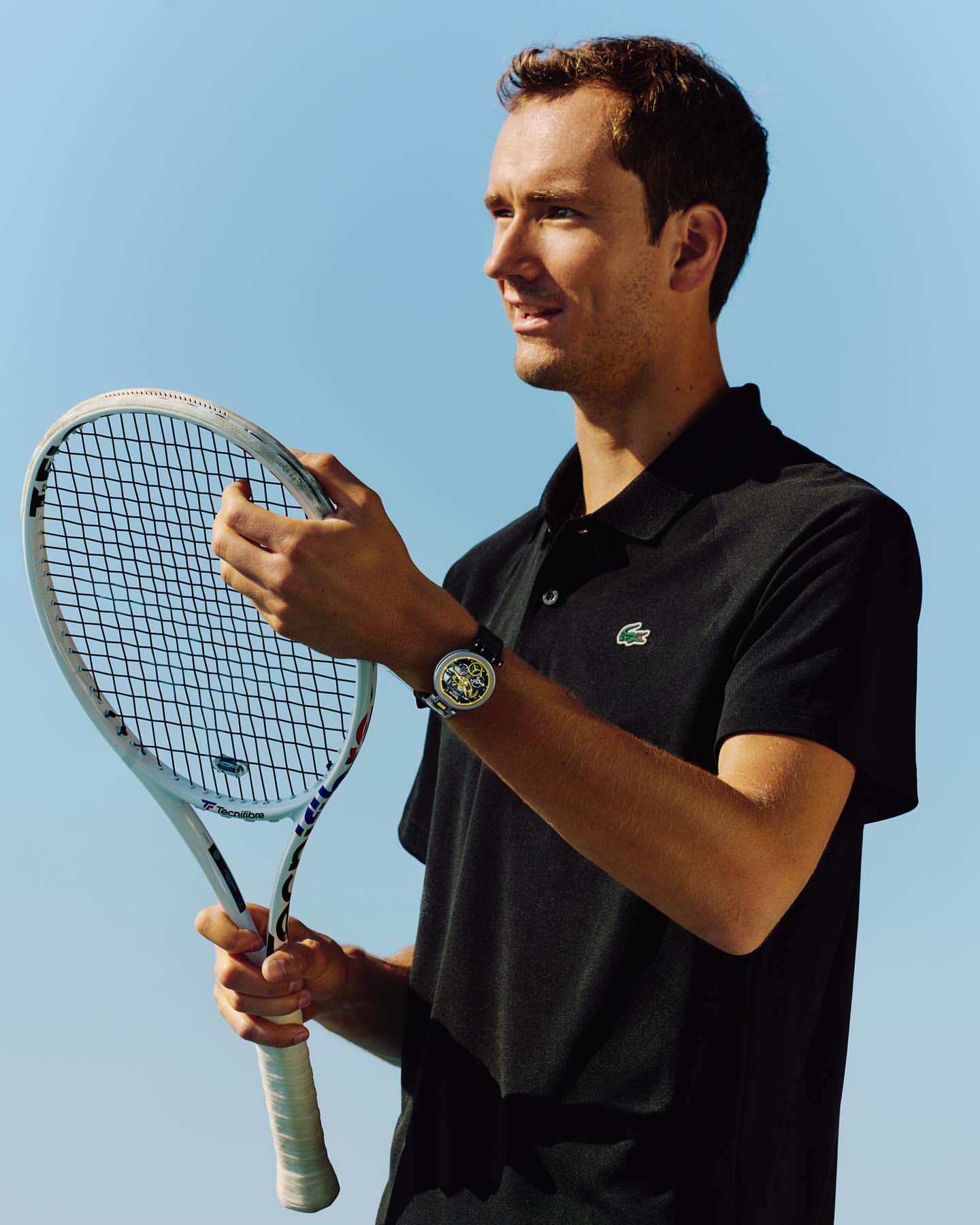Athletic male model poses outdoors, holding a white tennis racket and wearing a dark polo shirt with a recognizable Lacoste logo.