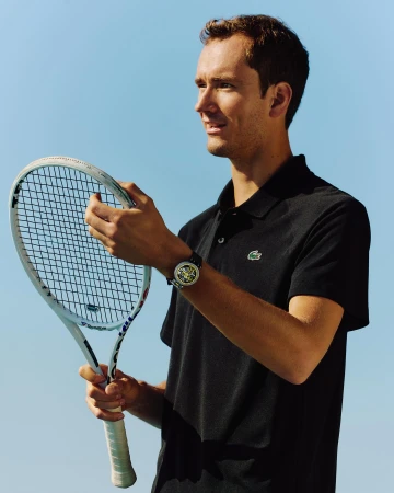 Athletic male model poses outdoors, holding a white tennis racket and wearing a dark polo shirt with a recognizable Lacoste logo.