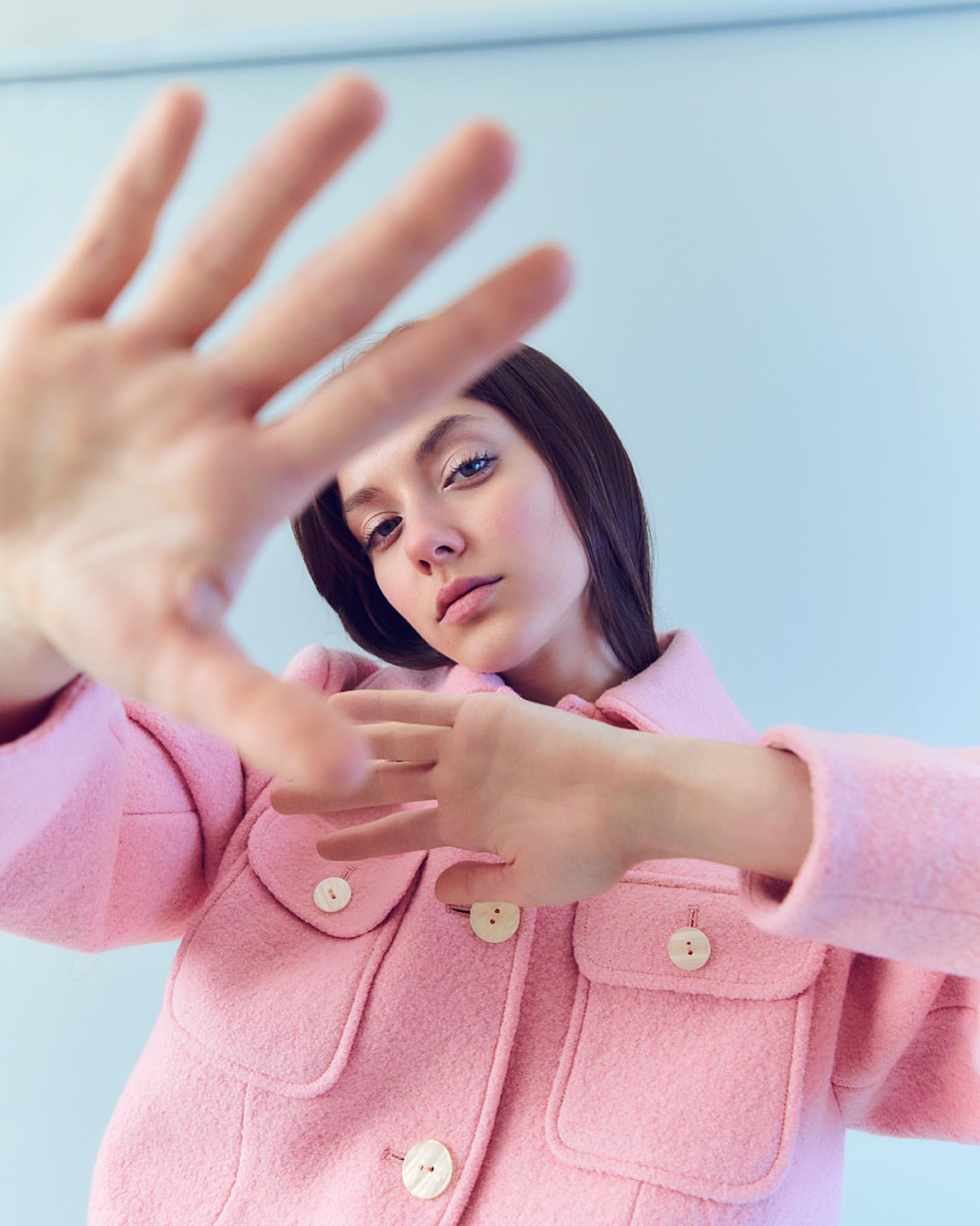 Stylish young woman in a pink jacket poses with her hands framing the camera, set against a light blue background in a fashion setting.