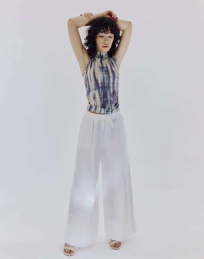 A young woman with curly dark hair stands in a sleeveless tie-dye top and white palazzo pants against a bright white backdrop in a studio shot.
