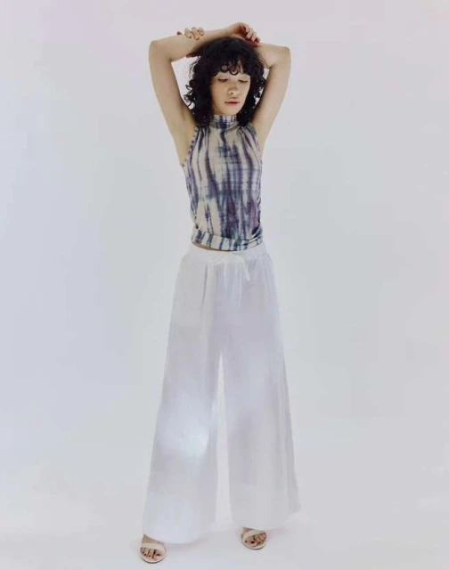 A young woman with curly dark hair stands in a sleeveless tie-dye top and white palazzo pants against a bright white backdrop in a studio shot.