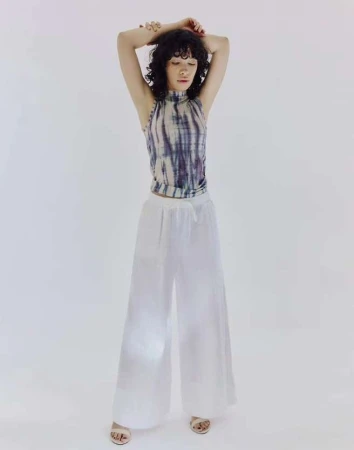 A young woman with curly dark hair stands in a sleeveless tie-dye top and white palazzo pants against a bright white backdrop in a studio shot.