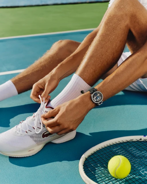Close-up of a person tying their white Lacoste tennis shoe, with a yellow tennis ball and tennis racket visible on a blue tennis court.