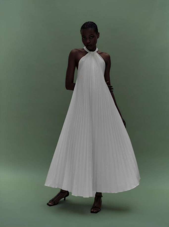 Elegant Black woman in a white pleated halter dress and sandals, posing in a minimalist studio setting with a green backdrop.