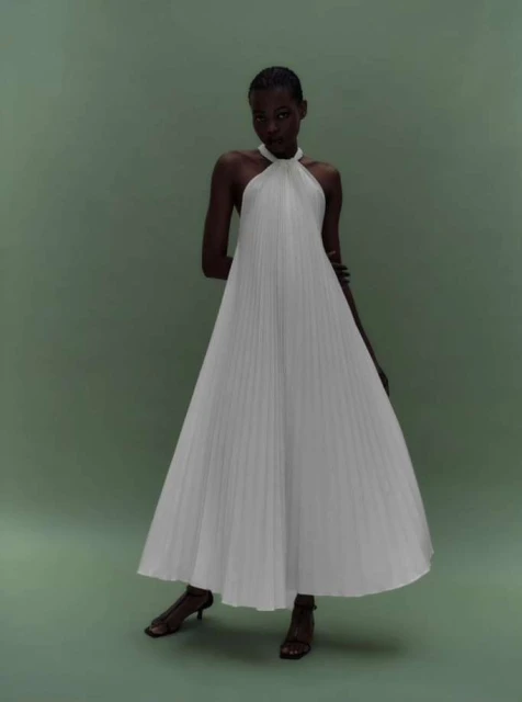 Elegant Black woman in a white pleated halter dress and sandals, posing in a minimalist studio setting with a green backdrop.
