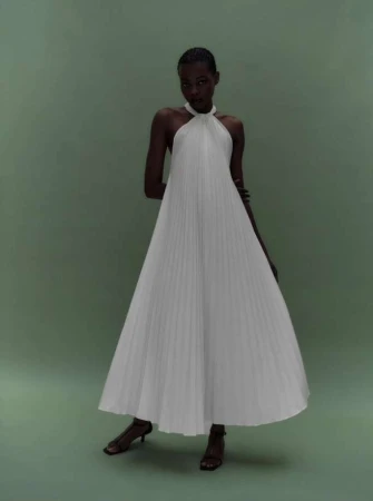 Elegant Black woman in a white pleated halter dress and sandals, posing in a minimalist studio setting with a green backdrop.