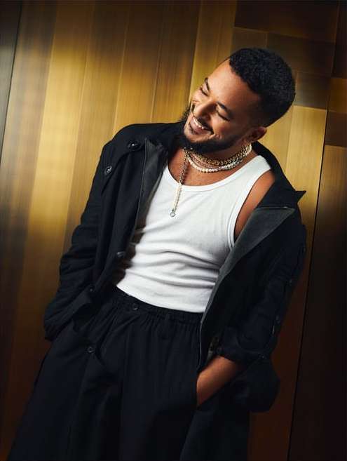 Smiling man in a white tank top, black coat, and black pants poses against a tan backdrop. He wears layered necklaces.