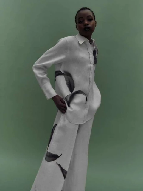Elegant African-American woman posing in a white linen shirt and pants with leaf prints against a light green background in a fashion shot.