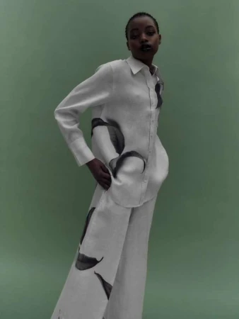 Elegant African-American woman posing in a white linen shirt and pants with leaf prints against a light green background in a fashion shot.