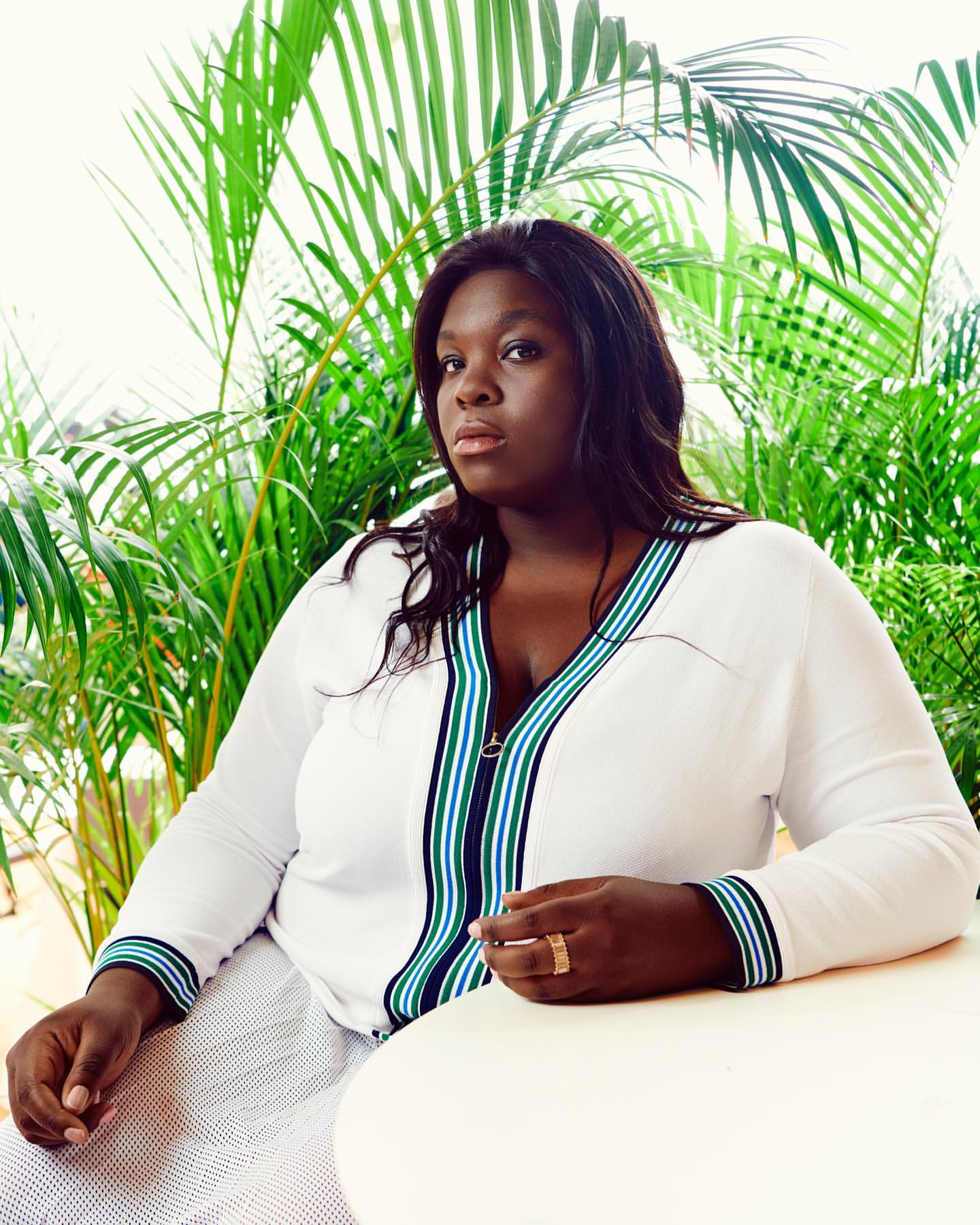 Portrait of an adult woman with dark hair, wearing a white blouse with green and blue stripes, posing in front of palm fronds.