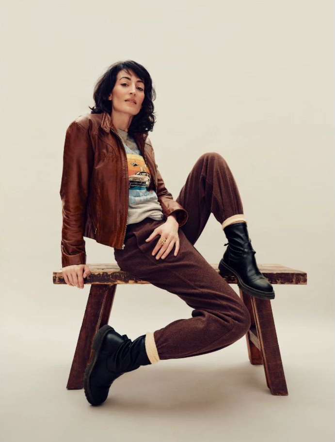 Woman in brown leather jacket and trousers sitting on a wooden bench against a neutral background