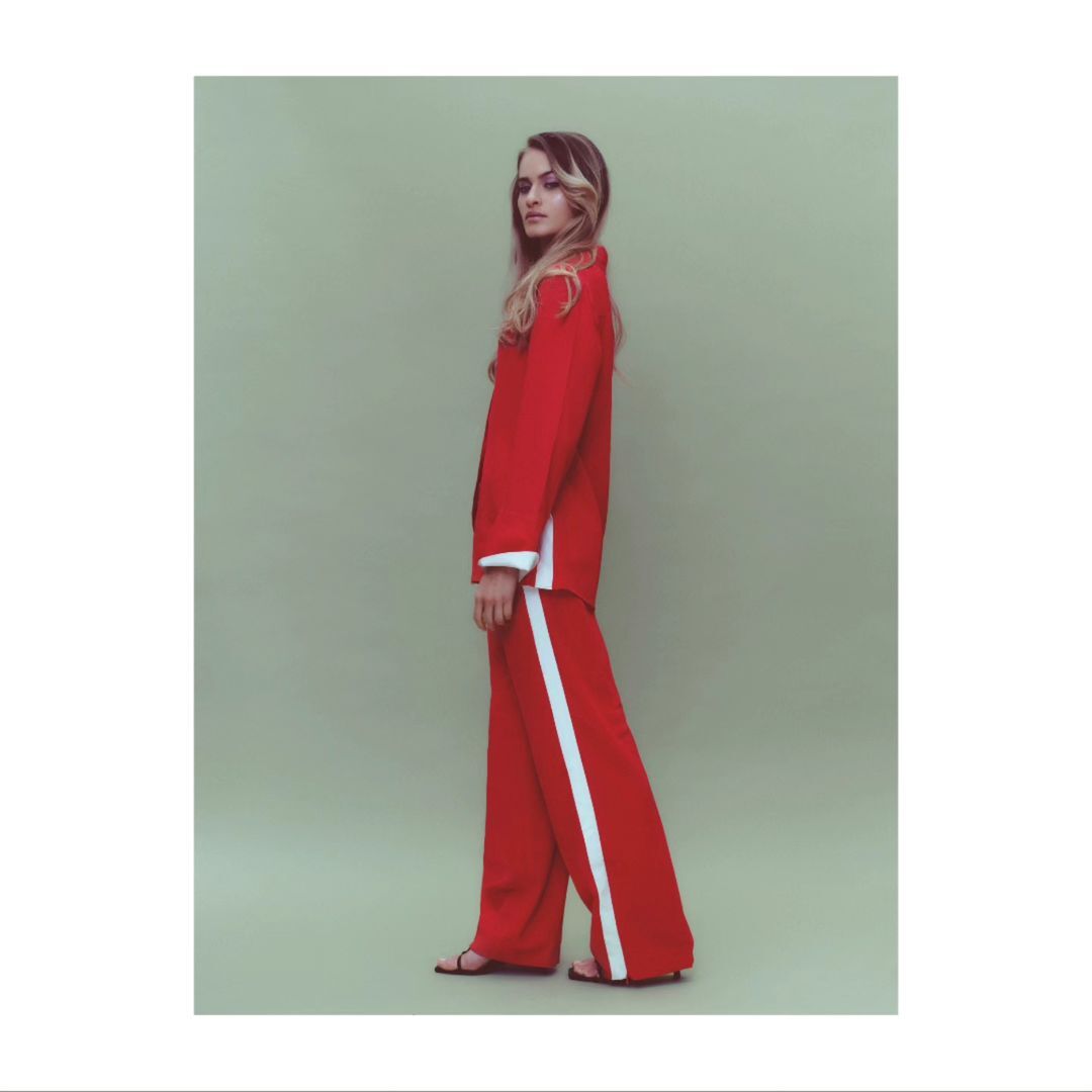 Fashion shot of a blond woman wearing a red suit with white stripes and sandals in a light green photo studio setting. Full-body, side view.