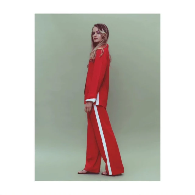 Fashion shot of a blond woman wearing a red suit with white stripes and sandals in a light green photo studio setting. Full-body, side view.
