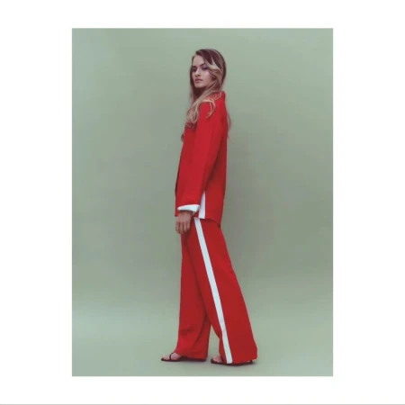 Fashion shot of a blond woman wearing a red suit with white stripes and sandals in a light green photo studio setting. Full-body, side view.