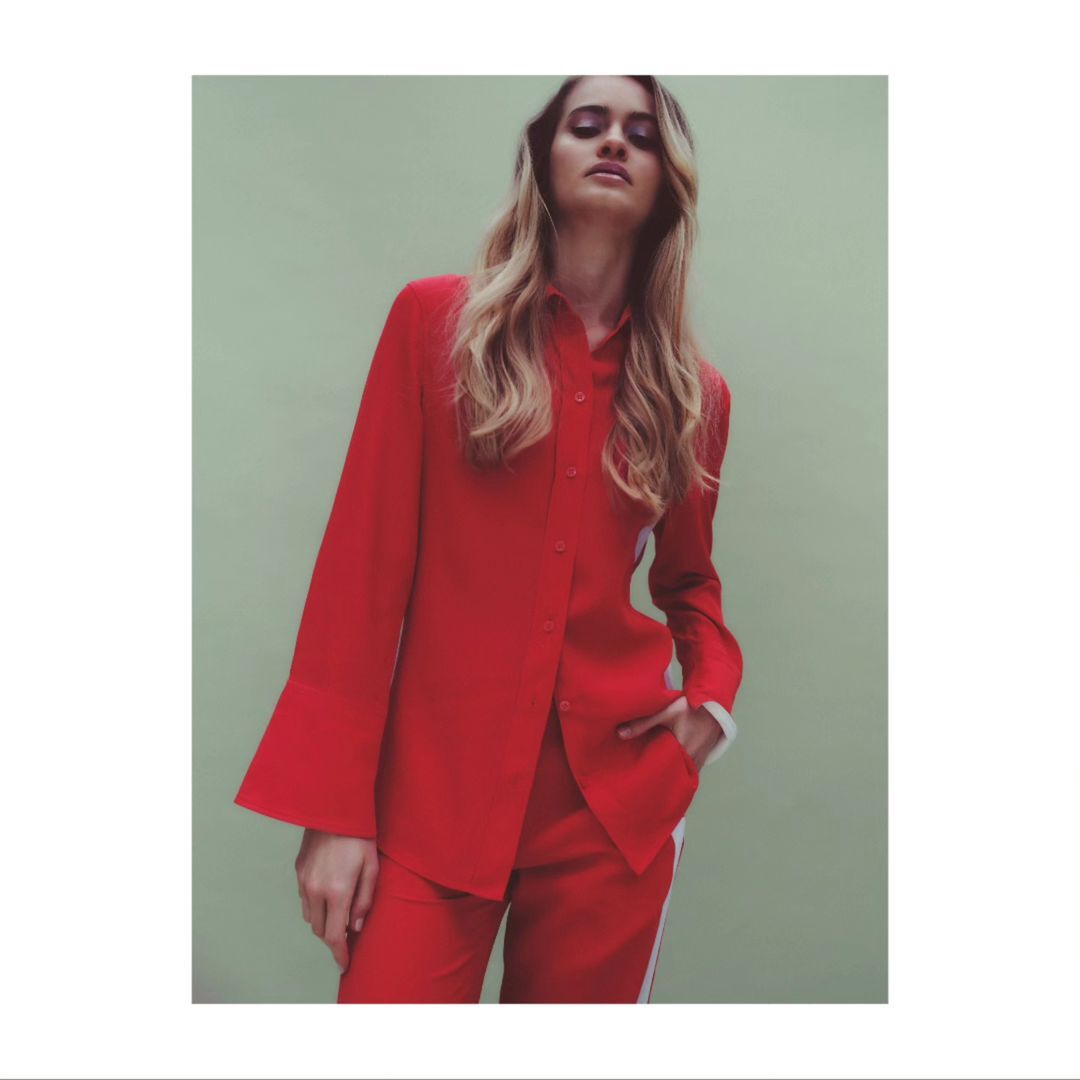 Woman in a red button-down shirt and red trousers posing against a muted green backdrop in Paris.