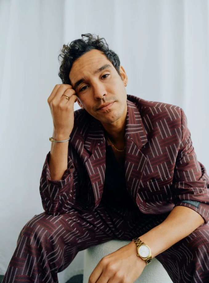 Stylish adult male in a maroon patterned suit sits indoors, glancing at the camera with a neutral expression in a medium studio shot.
