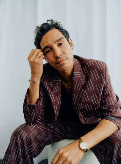 Stylish adult male in a maroon patterned suit sits indoors, glancing at the camera with a neutral expression in a medium studio shot.