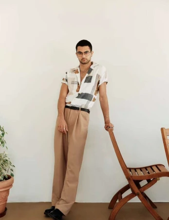 A stylish man with glasses, wearing a patterned shirt and tan pants, casually leans against a wooden chair indoors with a white wall background.