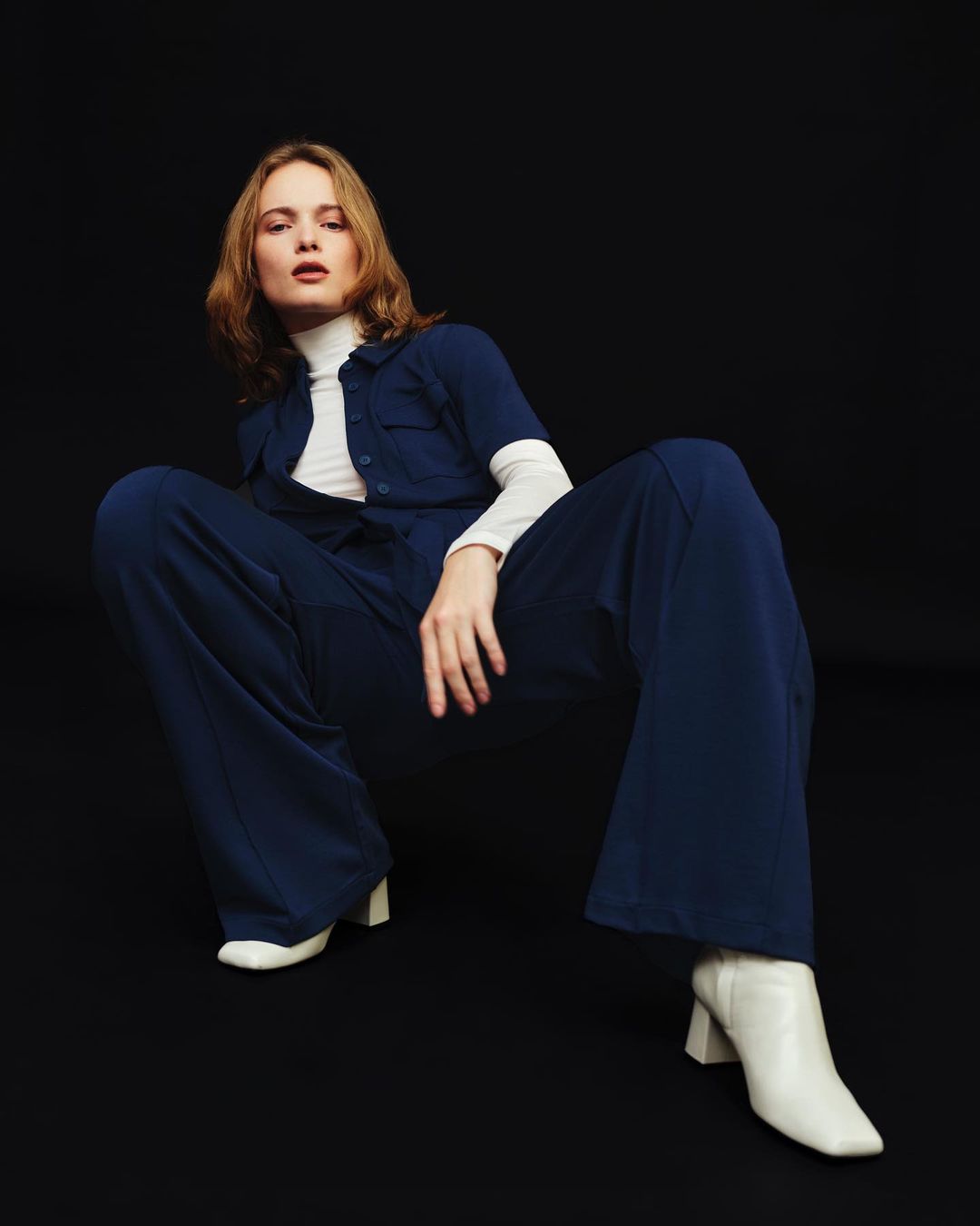 A young blonde woman poses in a navy blue jumpsuit and white boots against a dark background in a studio fashion shot.