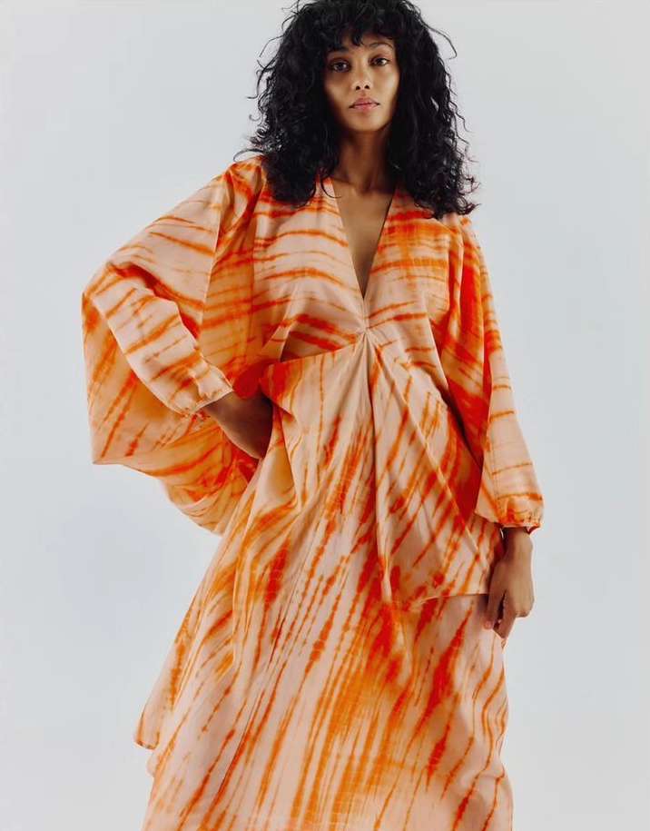 A woman with curly hair poses in a loose-fitting, tie-dye dress with a V-neck. The dress features a bright orange and peach color scheme.