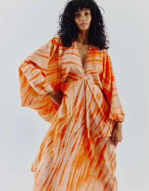 A woman with curly hair poses in a loose-fitting, tie-dye dress with a V-neck. The dress features a bright orange and peach color scheme.