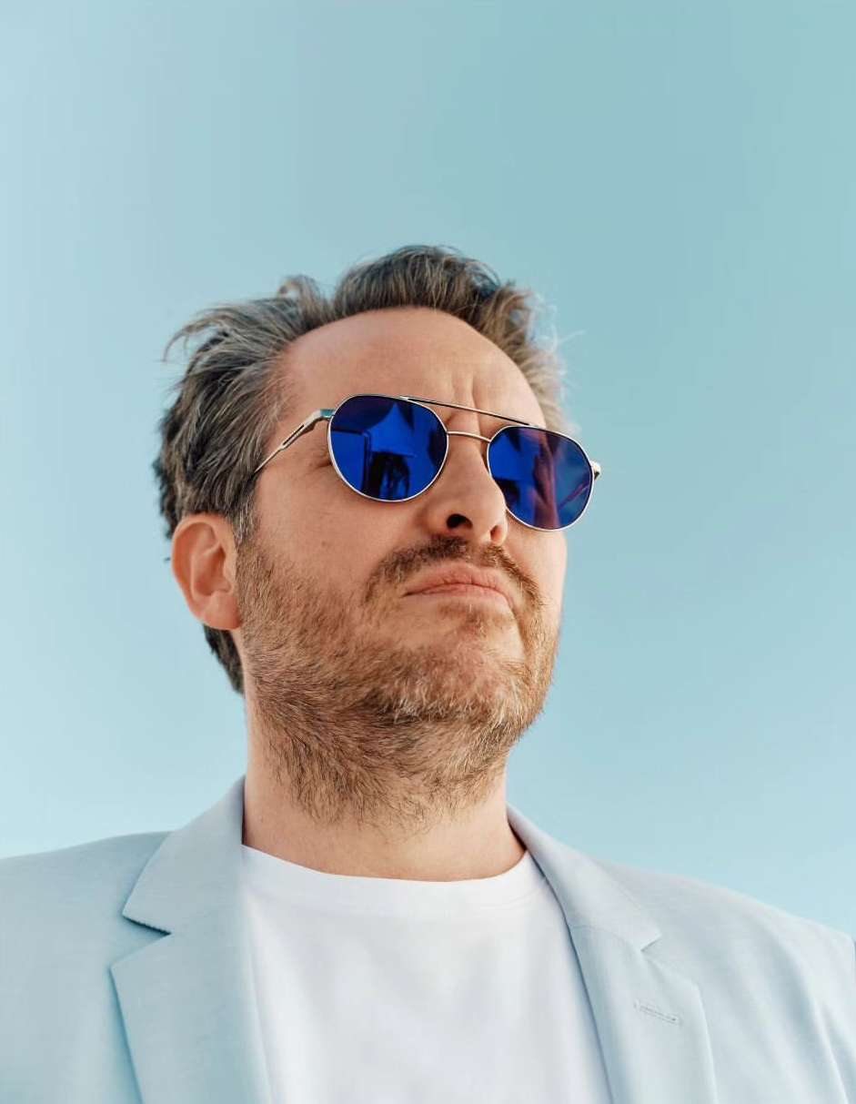 Close-up portrait of a man in a light-blue blazer and blue sunglasses against a matching light-blue background, gazing upwards.