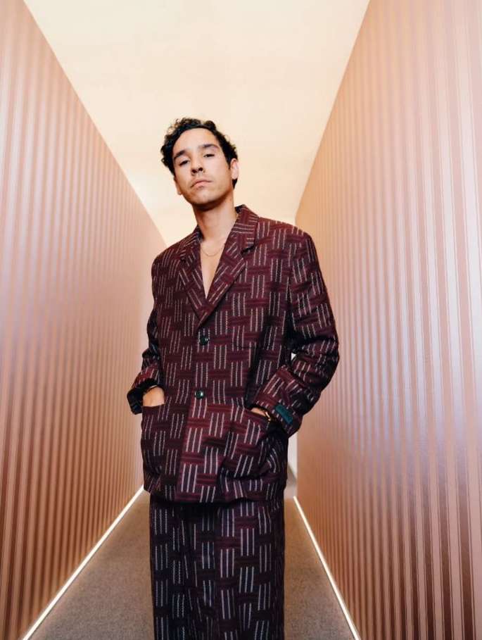 Man poses in a burgundy and white patterned suit in a hallway with peach colored striped walls and neutral lighting overhead.