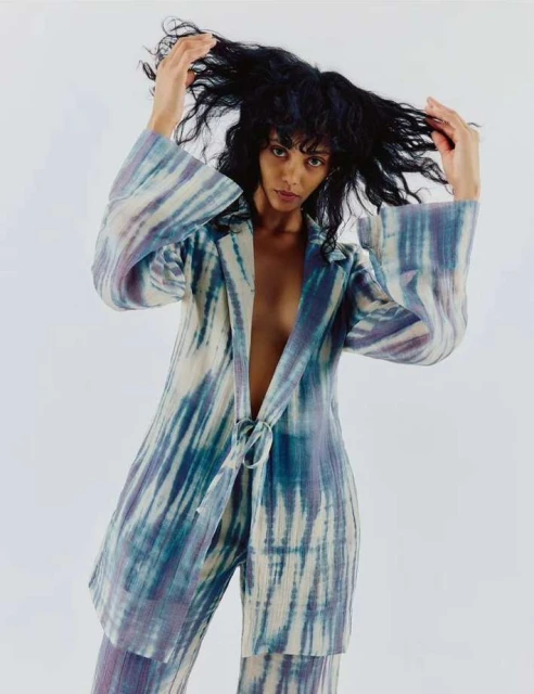 Woman in a tie-dye suit poses against a white backdrop, lifting her dark, curly hair with her hands in a fashion forward style.