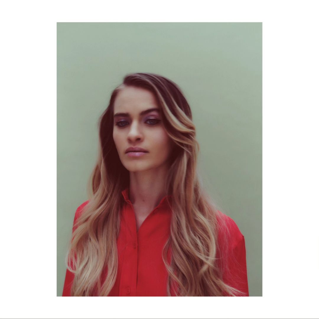 Portrait of an adult female in a red collared shirt against a green background. She has long, highlighted blonde hair with dark roots.