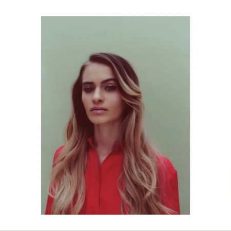Portrait of an adult female in a red collared shirt against a green background. She has long, highlighted blonde hair with dark roots.