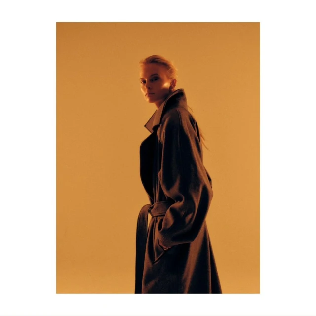 Stylish woman poses in studio wearing a long belted coat against a vibrant golden backdrop, showcasing fashion and elegant outerwear.