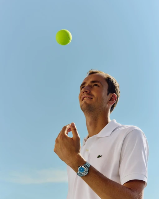 A tennis player in a white polo shirt looks up at a tennis ball against a blue sky, photographed from a low angle in natural light.