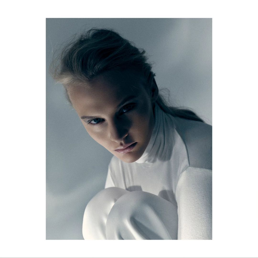 Portrait of a blonde woman with fair skin wearing a white turtleneck and pants, seated in a studio with soft, cool lighting.