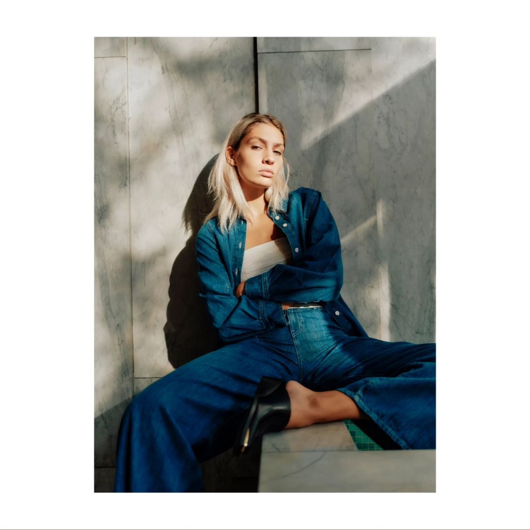 Blonde woman in denim outfit poses sitting against a marble wall with shadows in a fashion shot with natural lighting indoors.