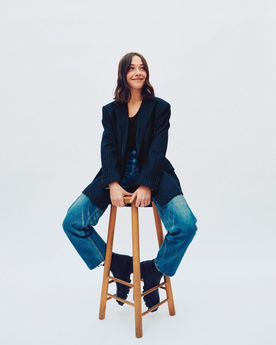 A young woman with wavy brown hair sits on a wooden stool, wearing a pinstripe blazer, black top, and blue jeans, with a white background.