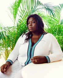Black woman with long dark hair wearing a white zip cardigan with striped trim seated near lush green palm fronds.