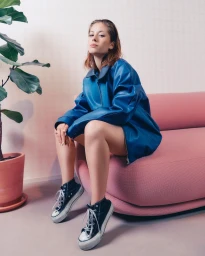 Woman in an oversized blue leather jacket sitting on a pink sofa wearing black high-top sneakers
