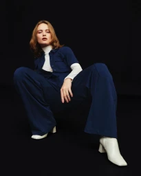 A female model in a blue utility jumpsuit over a white turtleneck and white boots squatting against a black background.