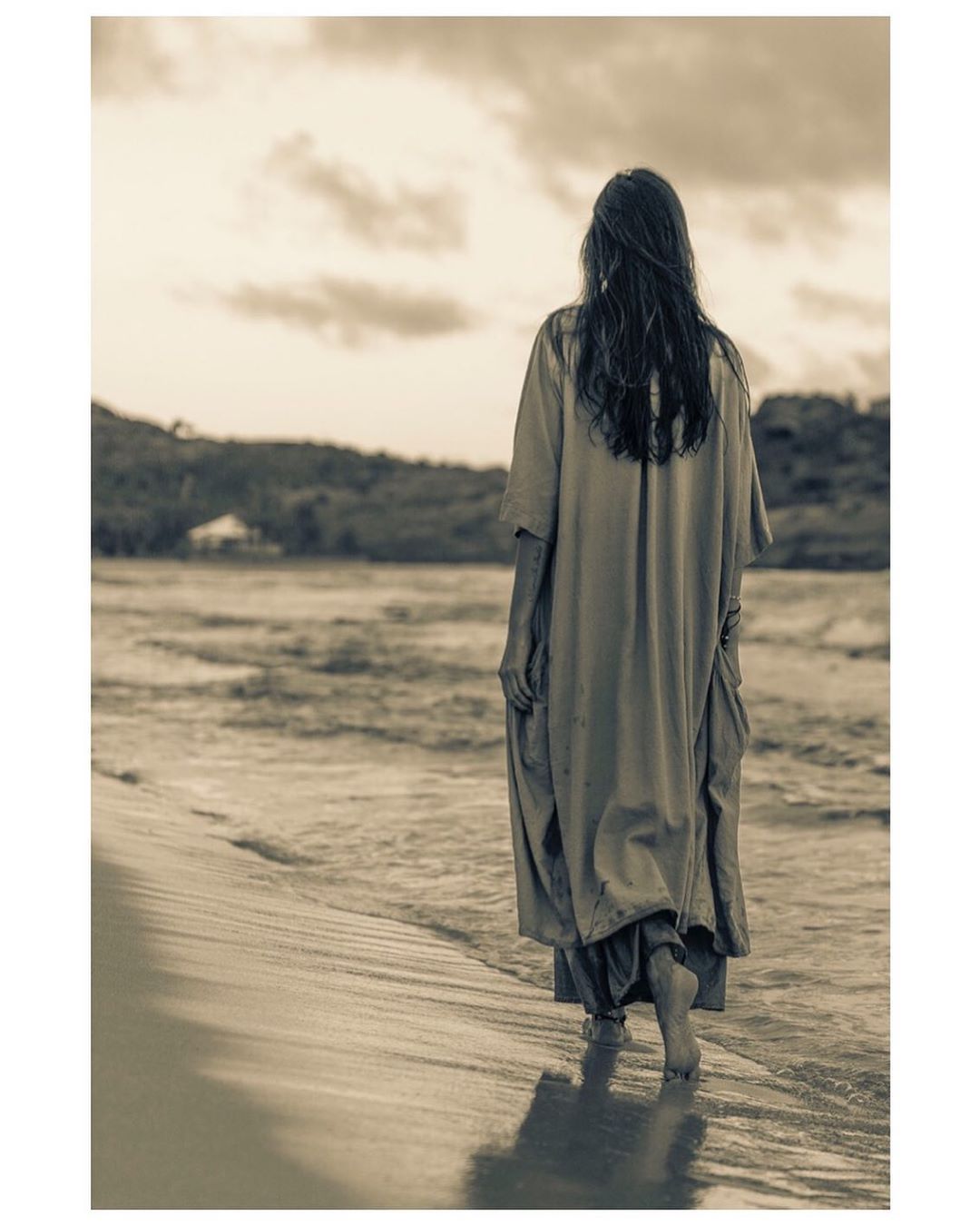 Back view of woman in flowing dress walking on sandy beach at sunset, with gentle waves and distant hills, in a sepia toned photograph.