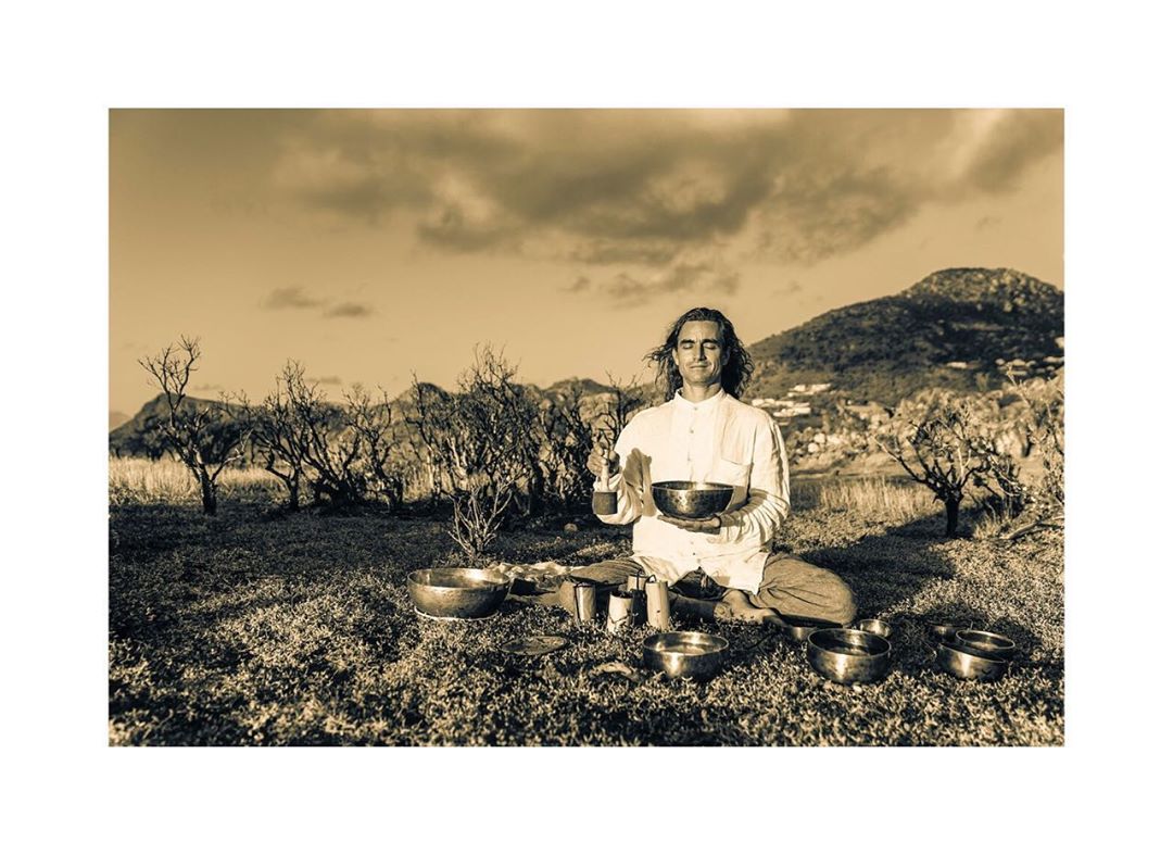 Adult male sits in nature, holding singing bowl. Bowls, mountains, and cloudy sky fill the background in a sepia-toned outdoor scene.