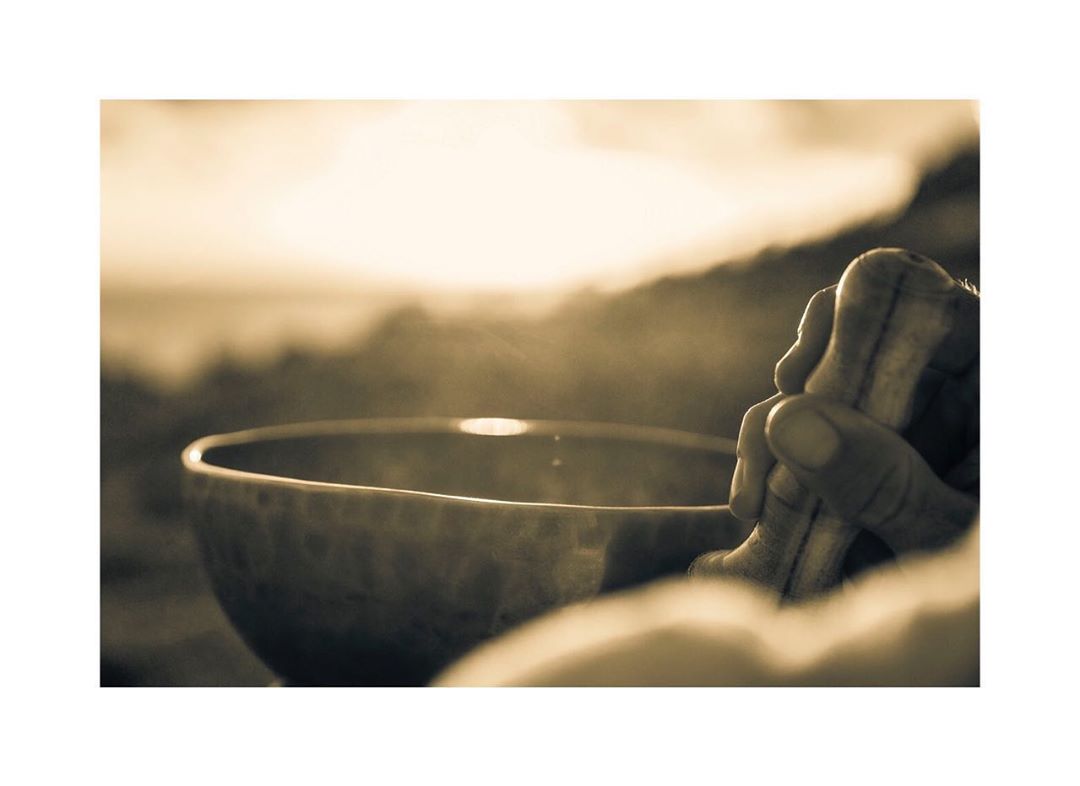 Close-up of a person's hands holding a mallet, poised over a singing bowl, with a blurred outdoor background in a sepia-toned image.