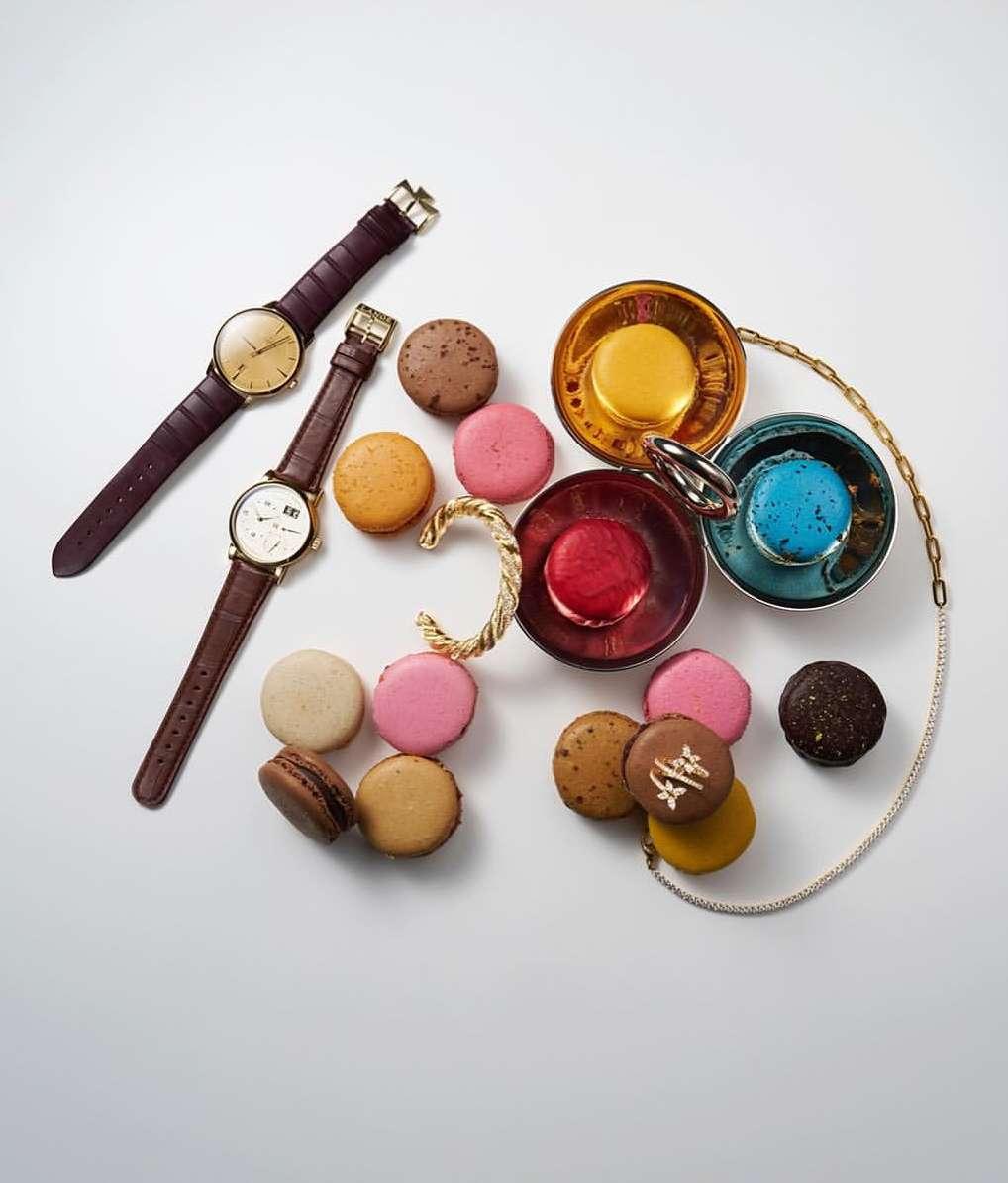 Overhead view of two wristwatches, macarons in different colors, a gold cuff, and a gold chain on a white background. Food and watches.