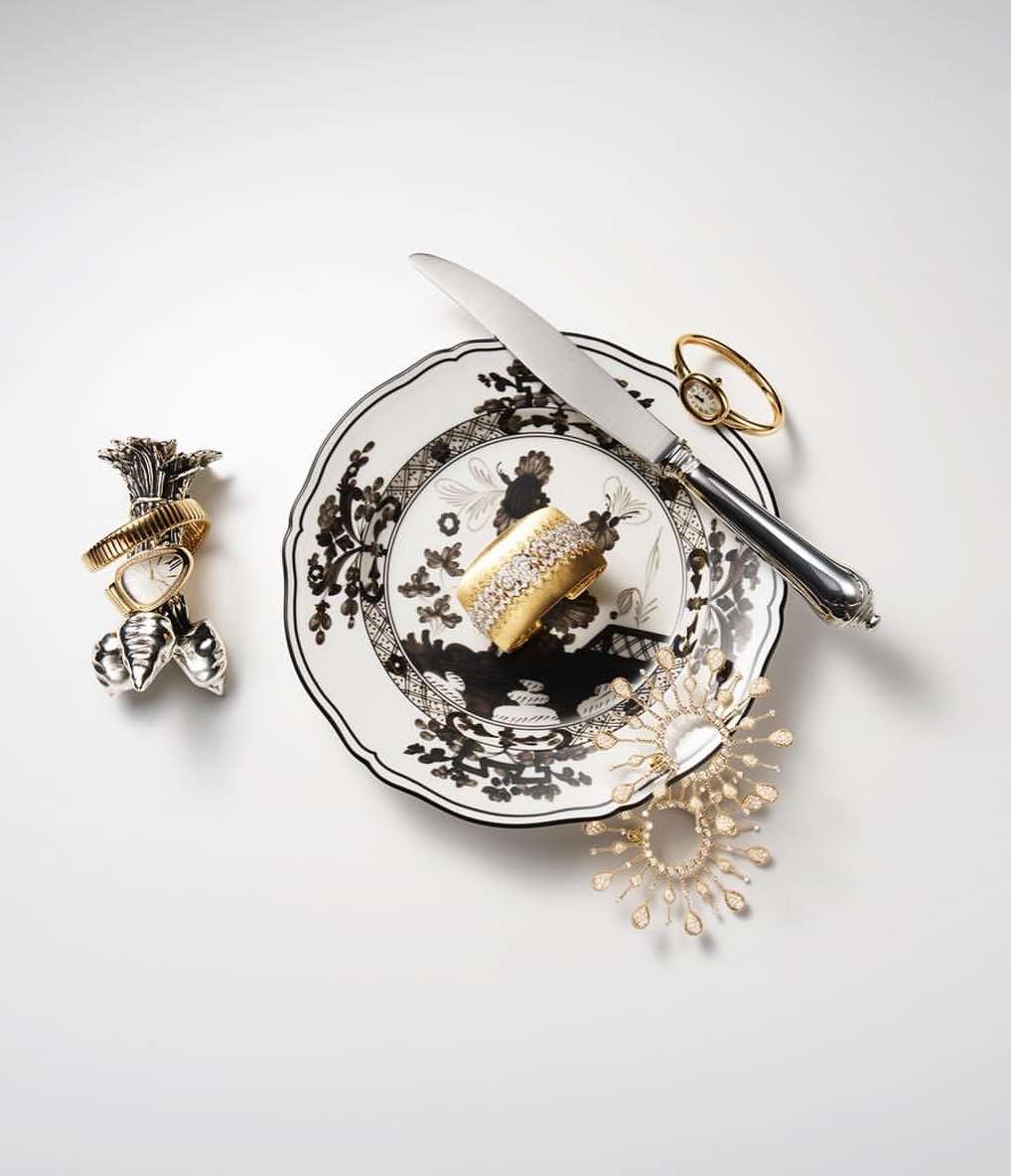 Still life of a black and white patterned plate with a gold cuff, a knife, and other jeweled items on a white backdrop.