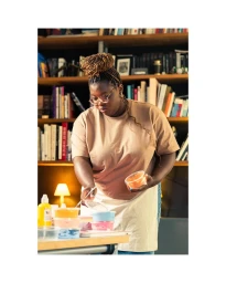 Black woman wearing glasses and a beige t shirt with an apron working at a desk filled with small plastic containers