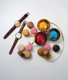 Overhead still life of colorful macarons, two luxury wristwatches, and gold jewelry on a white surface.